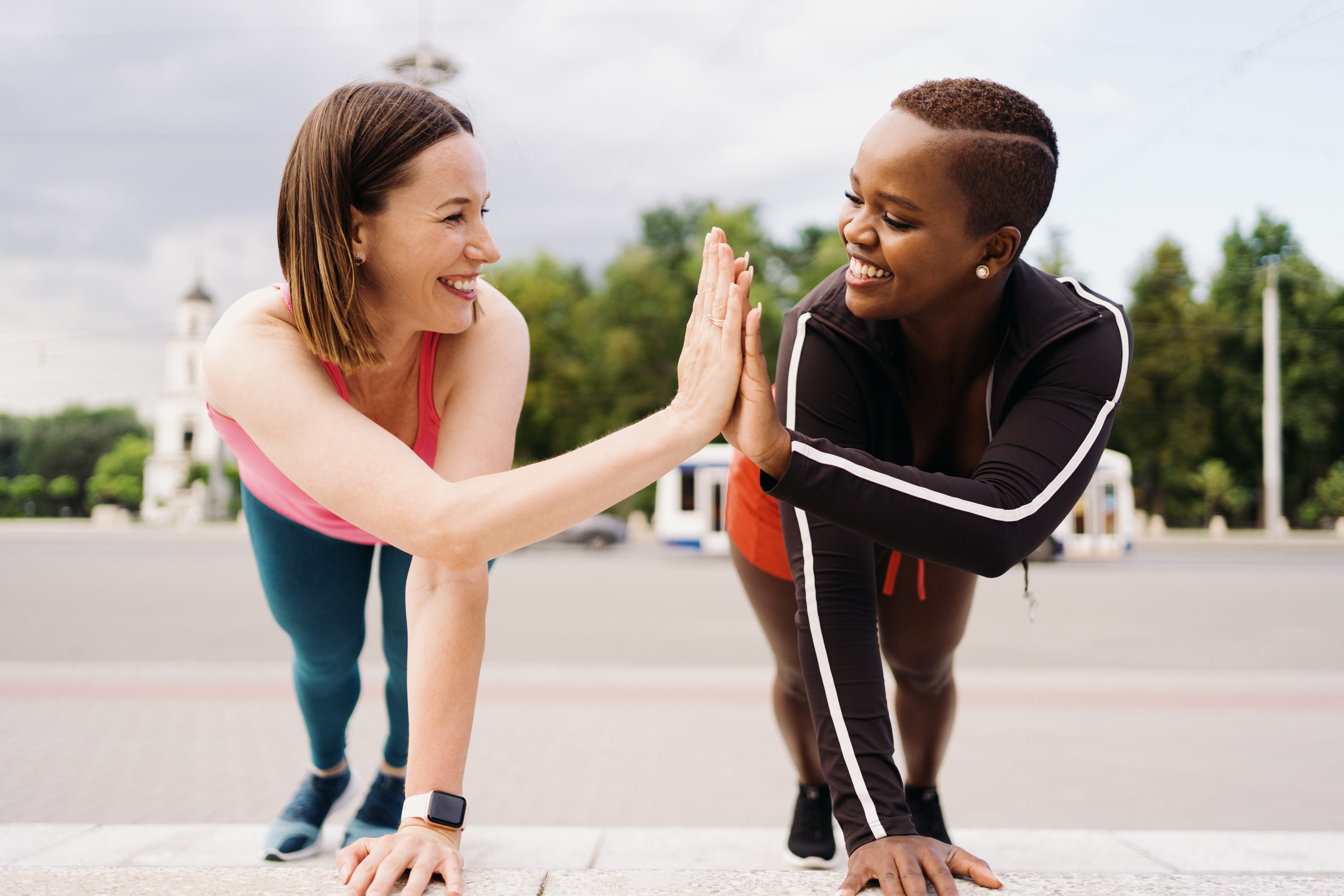 Two smiling diverse young woman in Athletic Workout Clothes are Doing a Plank Exercise working out together in city square.